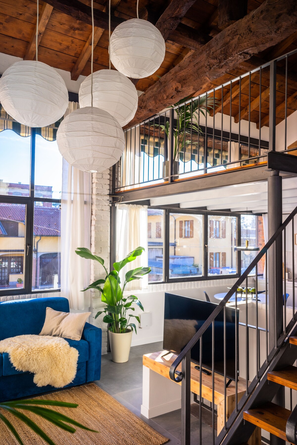 A bright living area features a blue sofa adorned with a soft throw. Hanging paper lanterns provide ambient lighting, while large windows allow natural light to enter. A green plant adds a touch of nature, and a wooden staircase leads to the upper level.