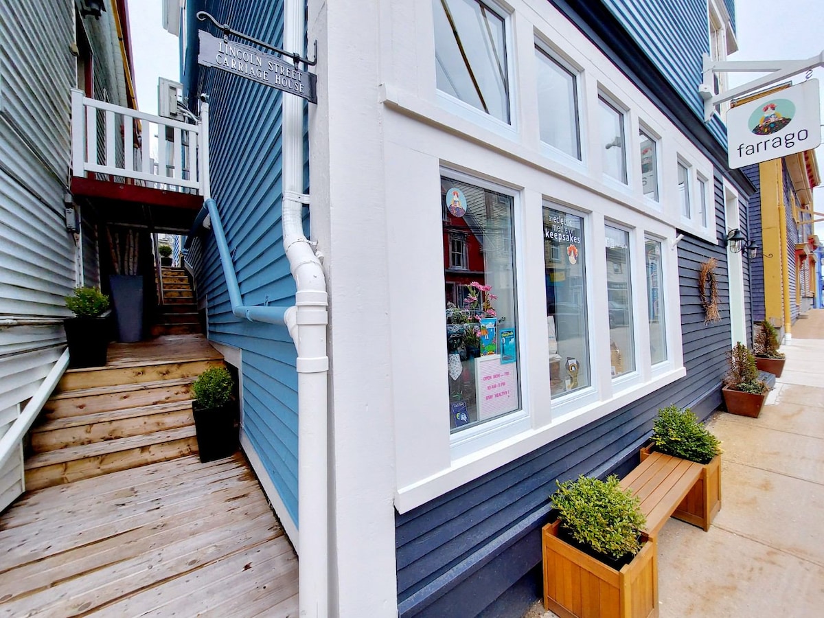 The image shows the exterior of the Lincoln Street Carriage House, featuring a charming blue facade with large windows. A wooden staircase leads to a private entrance, while planters with greenery are positioned alongside a bench by the entrance, enhancing the inviting appearance.