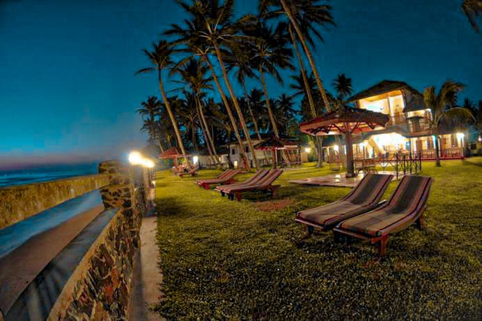 A spacious garden area is illuminated by soft lighting, surrounded by tall palm trees. Comfortable lounge chairs are arranged neatly on the grassy lawn, offering a view of the ocean. A stone wall borders the property, with torches providing a warm glow in the evening.