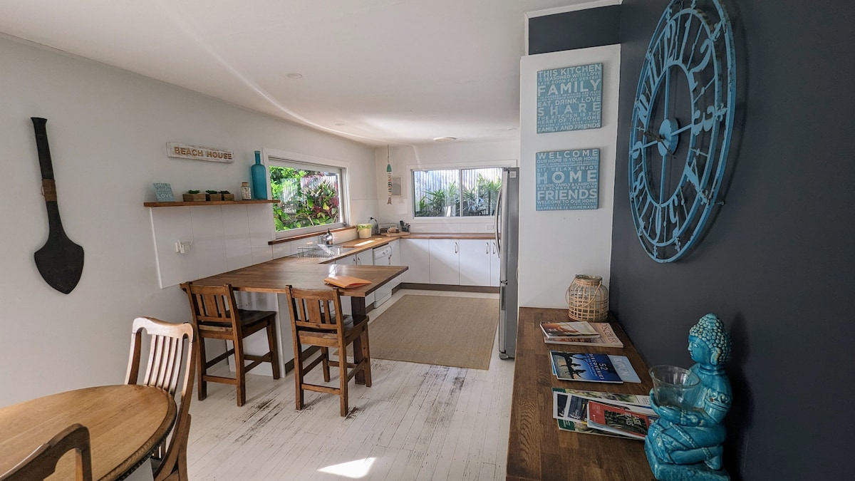 The spacious kitchen area features a large window allowing natural light to illuminate the space. A wooden dining table with chairs is positioned near the entrance, complemented by a decorative bicycle clock and shelves showcasing books and decor. Light-colored flooring enhances the open feel.