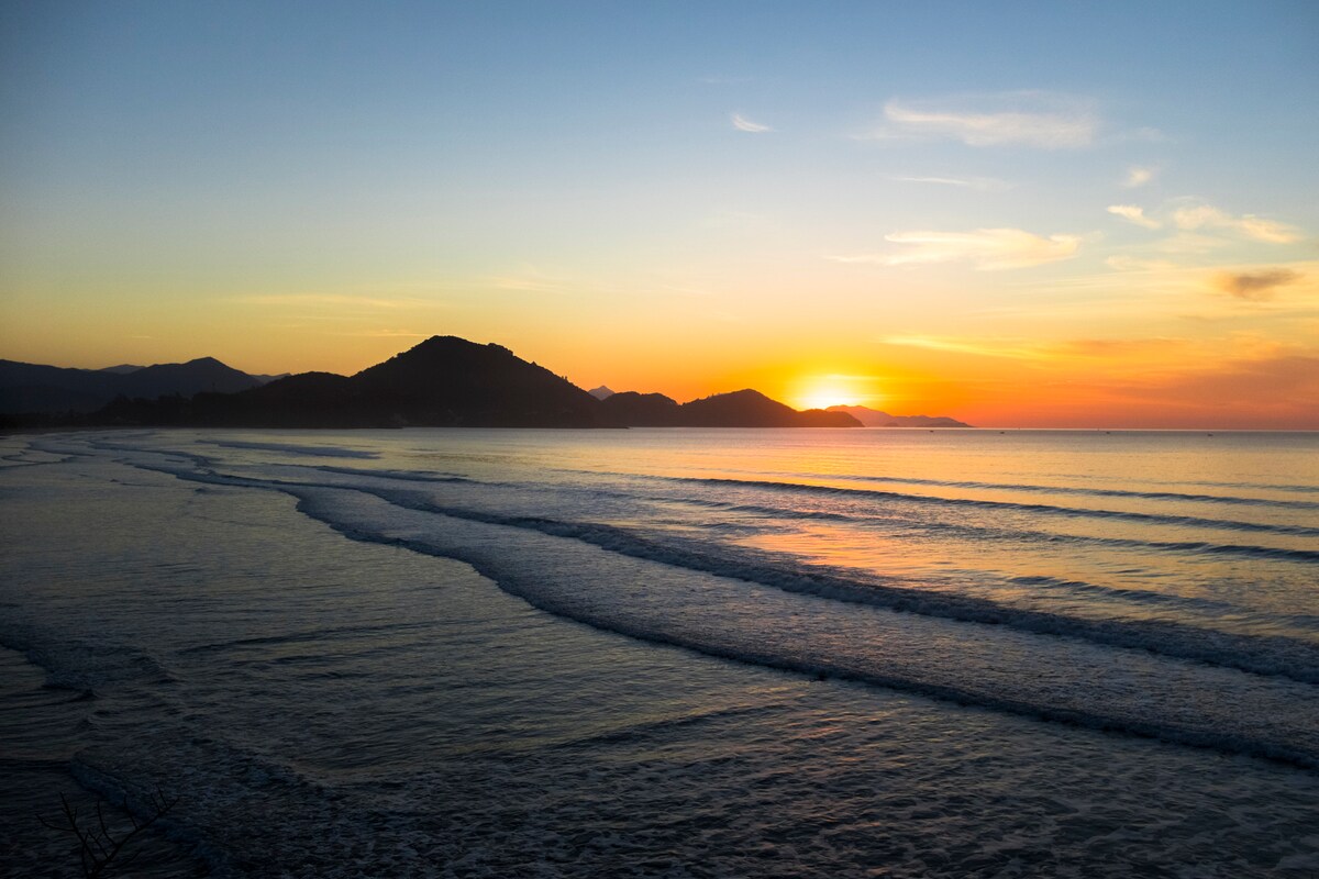 A serene coastal view captures the ocean waves gently lapping at the shore under a vibrant sunrise. Mountains silhouette against the horizon, while the sky transitions through soft hues of orange and blue, signaling the start of a new day.