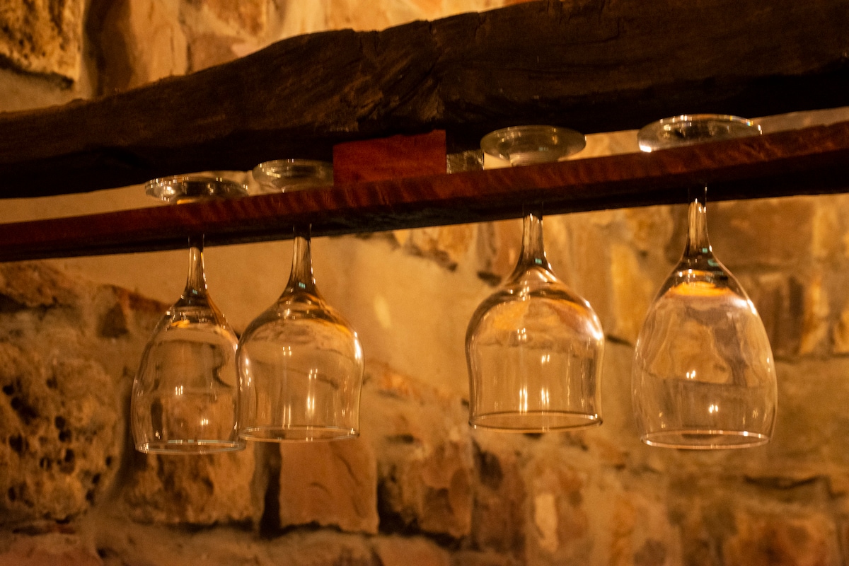 A wooden beam supports several hanging glass cups, showcasing their clear, reflective surfaces. The rustic stone wall in the background adds texture to the setting, enhancing the cabin's natural ambiance.