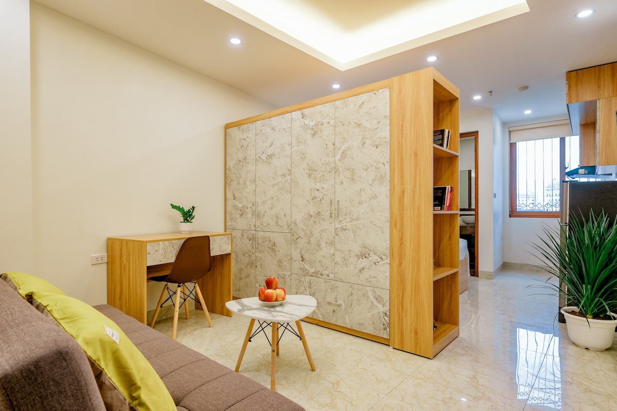 A modern living area is designed with light-colored marble flooring and wood accents. A compact desk and chair are positioned next to a decorative bookshelf. A cozy sofa and a small table are visible, complemented by a touch of greenery from a potted plant.