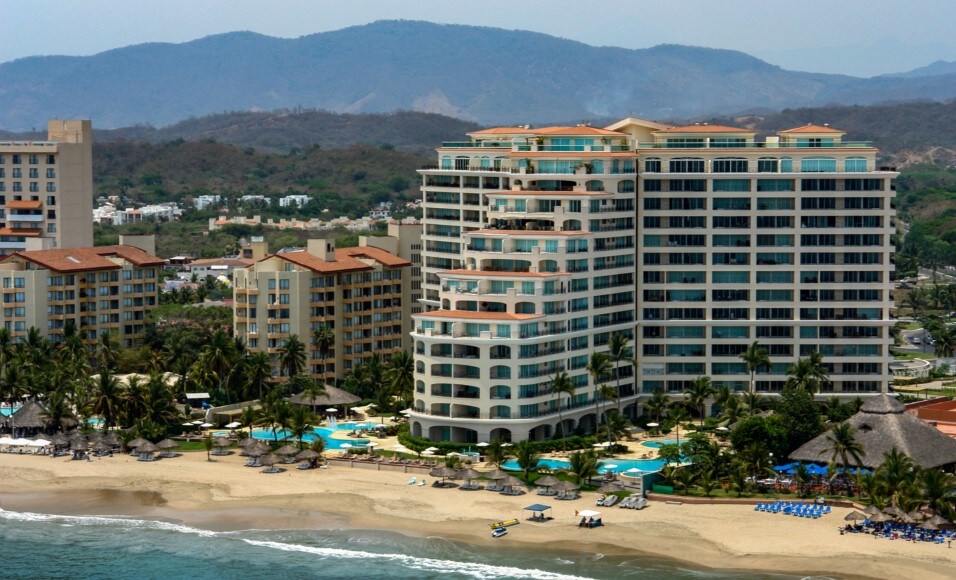 The beachfront building rises elegantly, surrounded by swaying palm trees and sandy shores. Multiple swimming pools are visible, each set against the backdrop of the ocean. Mountains create a serene horizon, complementing the overall coastal scenery.