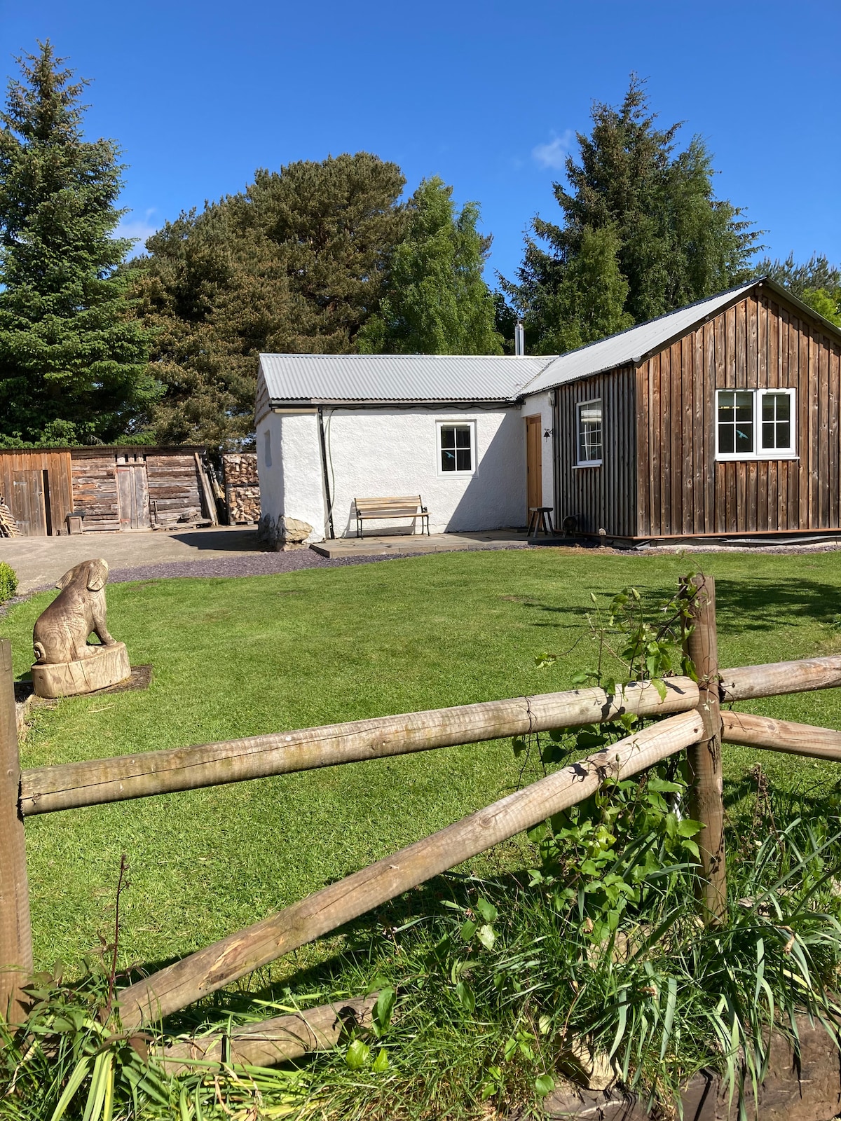 A charming cottage is nestled in a lush green landscape. The exterior features a combination of white and wooden cladding, with large windows allowing natural light. A well-maintained garden surrounds the property, complete with a wooden fence and a stone sculpture enhancing the tranquil setting.