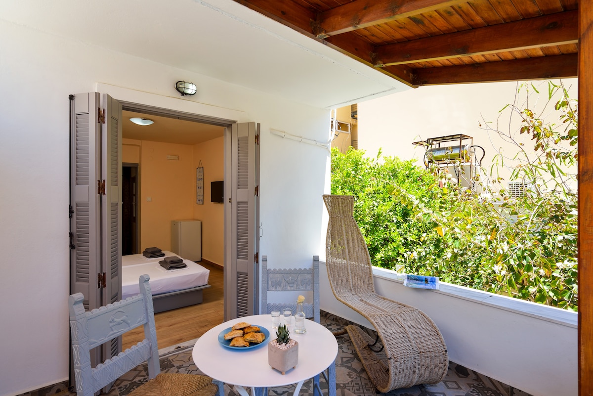 A private terrace is featured, furnished with a round table and two chairs. A cozy lounge chair is positioned nearby. Lush greenery is visible beyond the balcony, providing a serene backdrop. The entrance to the interior can be seen, with light-colored walls and minimal decor.