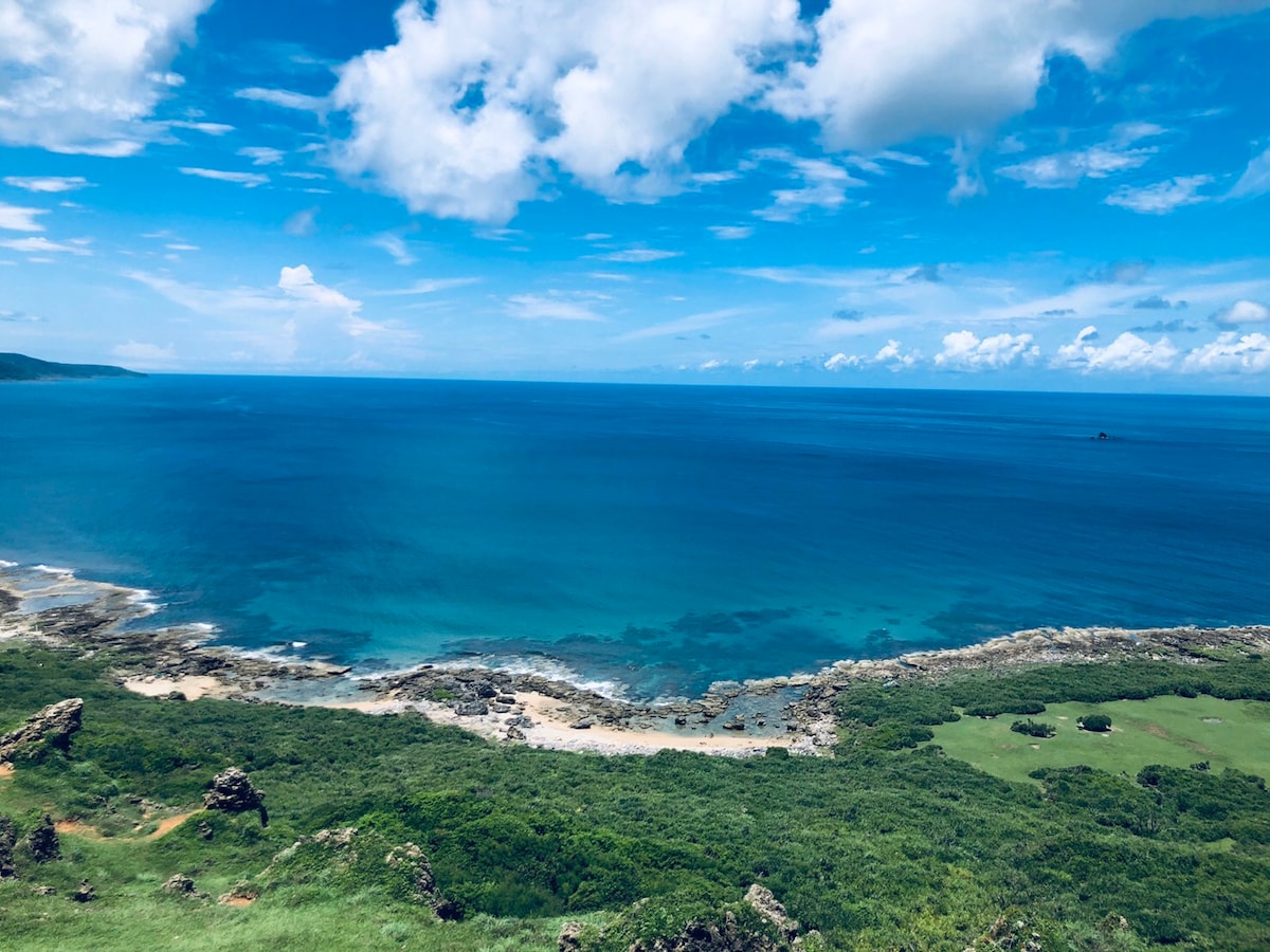 A wide view of a serene coastline is captured, showcasing clear blue waters lapping gently against rocky outcrops. Lush greenery lines the shore, contrasting with the sandy beach below. The sky is partly cloudy, allowing sunlight to reflect off the water's surface.