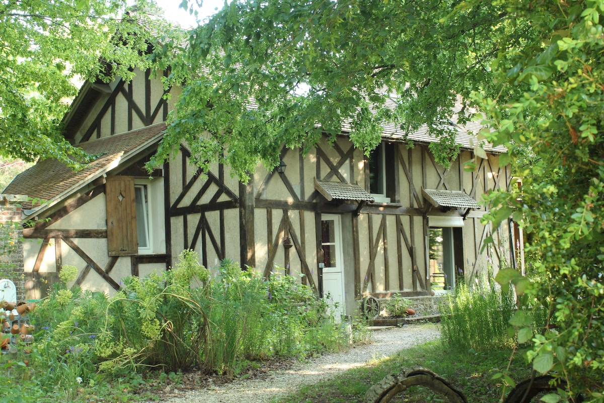 A charming traditional house is nestled among lush greenery, showcasing a combination of timber-framed architecture and natural surroundings. The exterior features multiple windows and wooden shutters, framed by vibrant foliage. A gravel path leads up to the entrance, creating a serene and inviting atmosphere.