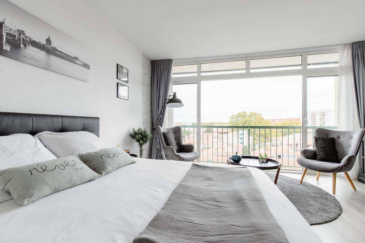 A well-lit bedroom features a spacious bed with light grey pillows displaying the word 'new.' A cozy seating area with two armchairs and a decorative table is positioned near a large window, providing a view of the outside landscape. Soft curtains frame the window, enhancing the natural light.
