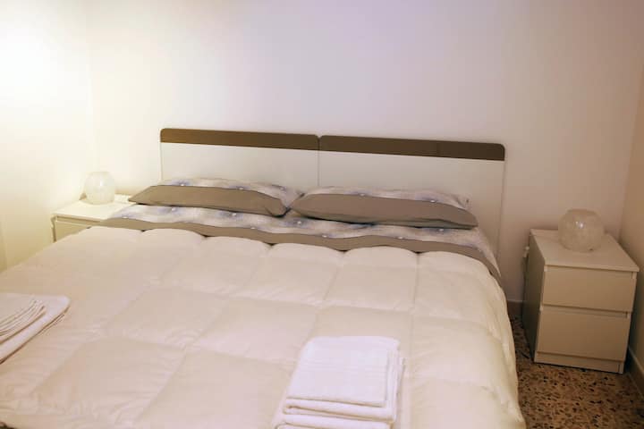 Eleganza minimalista: letto matrimoniale con cuscini grigi, lampada sul comodino e asciugamani pronti per gli ospiti.