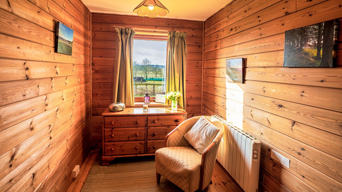A warm wooden interior is highlighted by natural light streaming through a window. A comfortable chair is positioned near a wooden dresser, and two framed pictures adorn the walls. Soft curtains gently frame the window, enhancing the calm ambiance.
