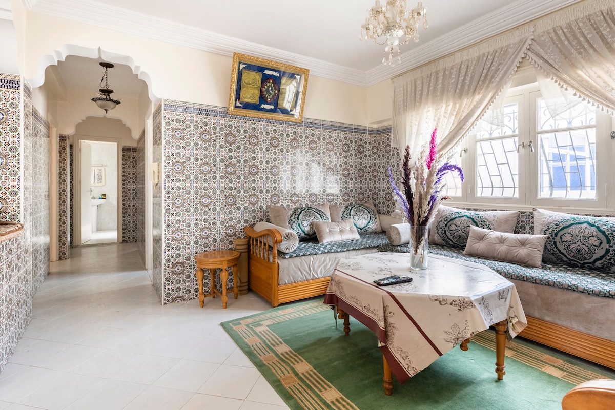 A spacious living area features intricate patterned tile walls and a large window allowing natural light. A comfortable seating area includes a traditional wooden bench adorned with decorative cushions, while a central table is set under an elegant chandelier.