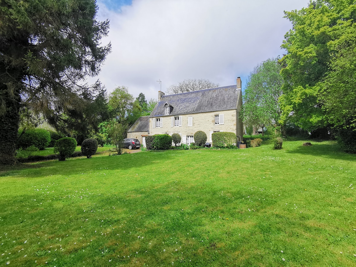 A traditional 18th-century family home is set within a spacious, lush green garden. The stone building features a gray roof and white window shutters, surrounded by well-maintained hedges. The expansive lawn invites outdoor activities in a tranquil, natural setting.