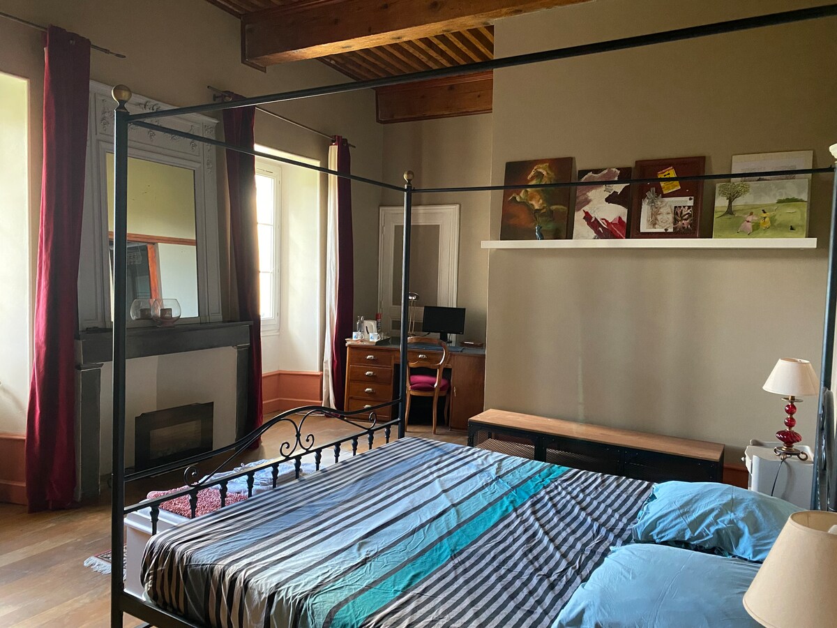 A spacious bedroom features a four-poster bed with a striped blanket and blue pillows. Large windows with red curtains allow natural light. A desk with a chair and wall-mounted artwork can be seen, along with a simple lamp and a wooden bench.