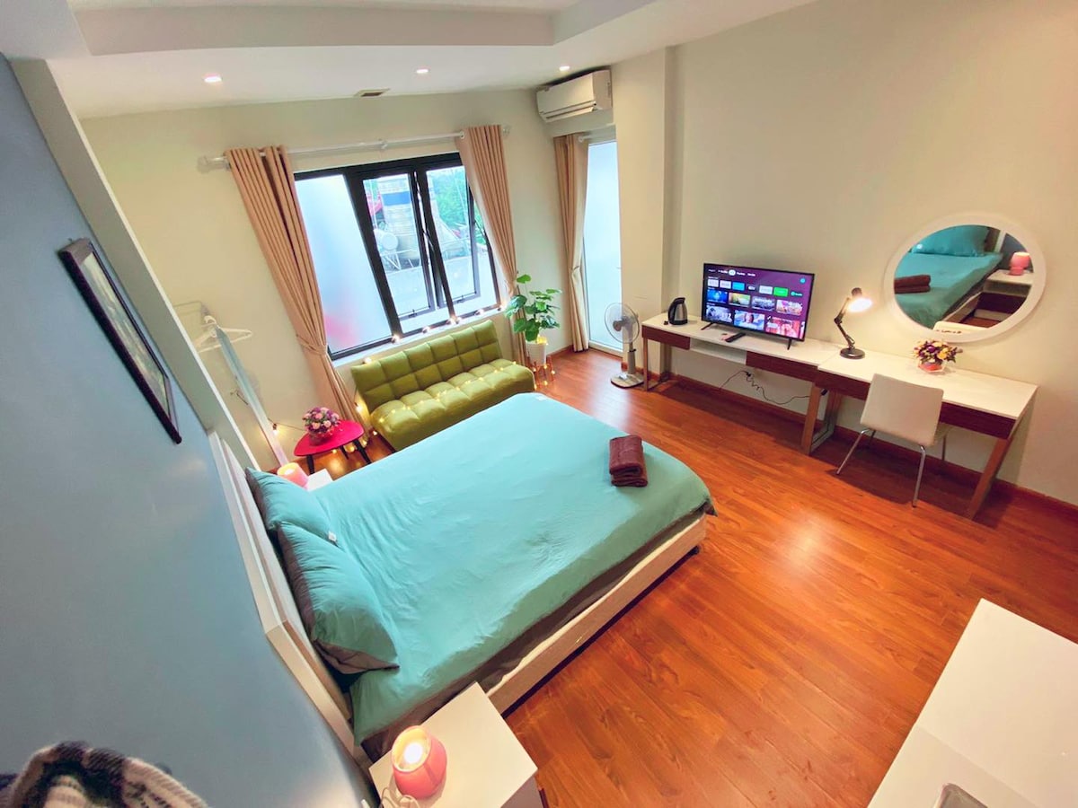 A modern studio space features a spacious bed with a soft blue coverlet, accompanied by a green sofa and a desk area with a mirror. A TV is visible on a nearby surface, while natural light filters in through large windows. Wood flooring complements the overall design.