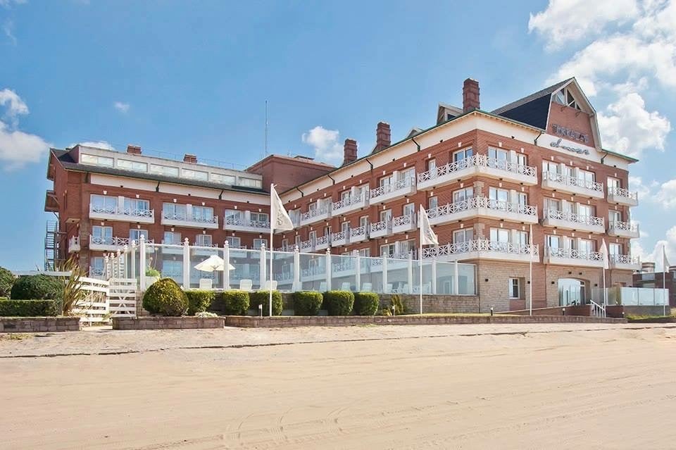 A three-story building with a brick facade is situated directly on the beach. Multiple balconies are visible, each adorned with white railings. Lush greenery and neatly trimmed hedges are present in the foreground, complemented by umbrellas offering shade along the sandy shore.