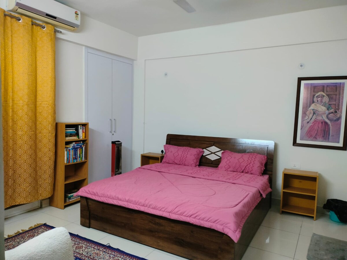 A bedroom is features a spacious bed with pink bedding. Two bedside tables are positioned on either side of the bed. A wooden bookshelf holds a selection of books. Bright curtains frame a window, allowing natural light to brighten the room.
