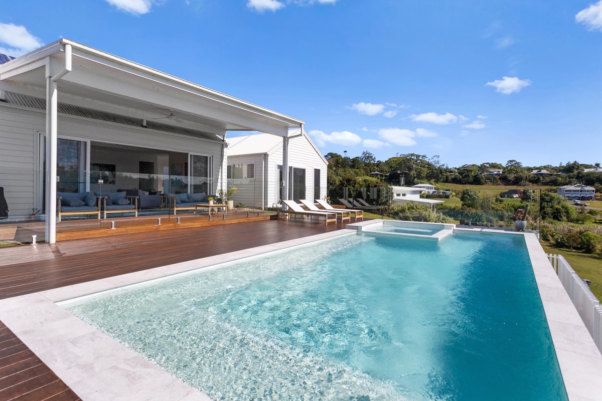 A spacious deck is shown overlooking a large magnesium pool and heated spa. Sun loungers are positioned beside the pool, with a covered outdoor living area visible in the background. The surrounding landscape features greenery and open space.