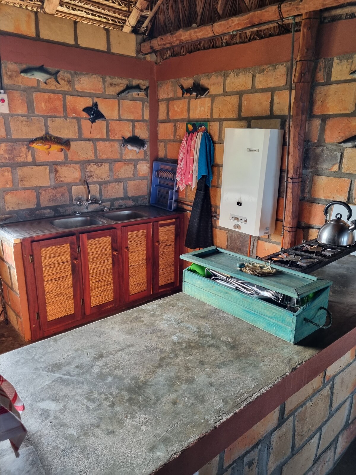 The kitchen area features a blend of brick and wooden cabinetry, equipped with a sink and a gas stove. Decorative fish are mounted on the walls, adding a touch of local charm. Cooking utensils are neatly arranged in a rustic green box on the counter.