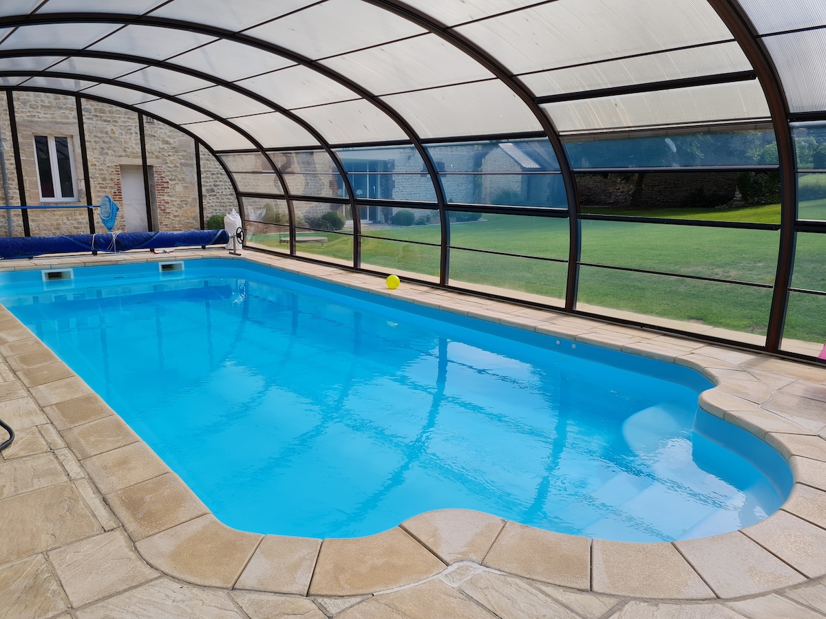 A covered and heated swimming pool is visible, featuring a smooth, curved shape and clear blue water. Surrounding the pool, a stone patio provides ample space for relaxation. The structure's transparent roof allows natural light to illuminate the area.