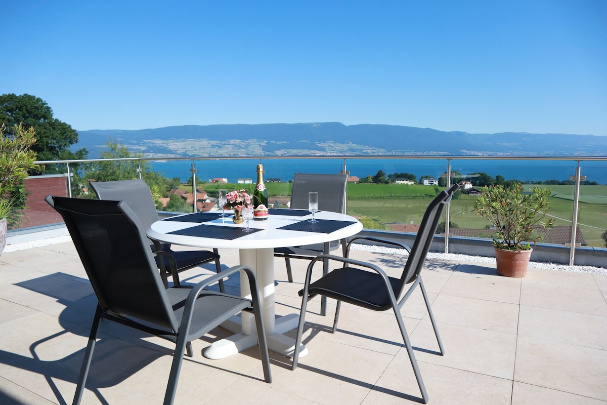 A terrace with a scenic view offers a round dining table surrounded by four gray chairs. The table is set with drinks and flowers, while a panoramic backdrop of lake and mountains extends into the distance under clear blue skies.