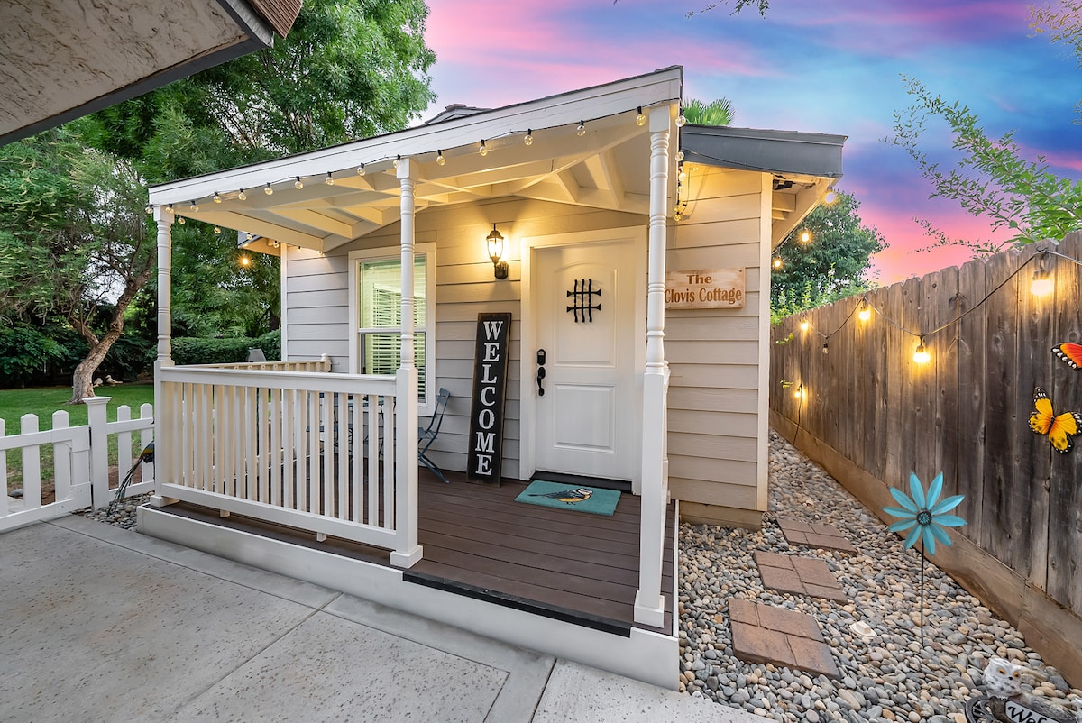 A welcoming porch features a light-colored wooden structure with a decorative sign displaying 'The Clovis Cottage.' String lights hang above, adding a warm glow to the inviting entryway. A small mat is placed in front of the door, which is surrounded by landscaped gravel and greenery.