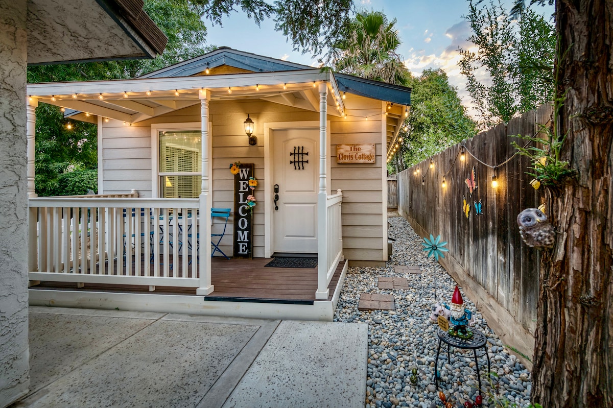The cottage exterior features a welcoming porch adorned with string lights, providing a cozy ambiance during twilight. A decorative sign displaying 'WELCOME' is placed prominently, alongside a blue chair set. The surrounding landscape includes gravel and greenery, enhancing the inviting atmosphere.