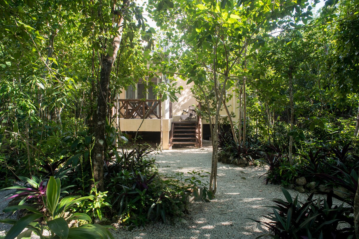 A bungalow is nestled among lush greenery, surrounded by a variety of tropical plants. The entrance features wooden steps leading up to a balcony with seating, inviting relaxation in a natural setting.