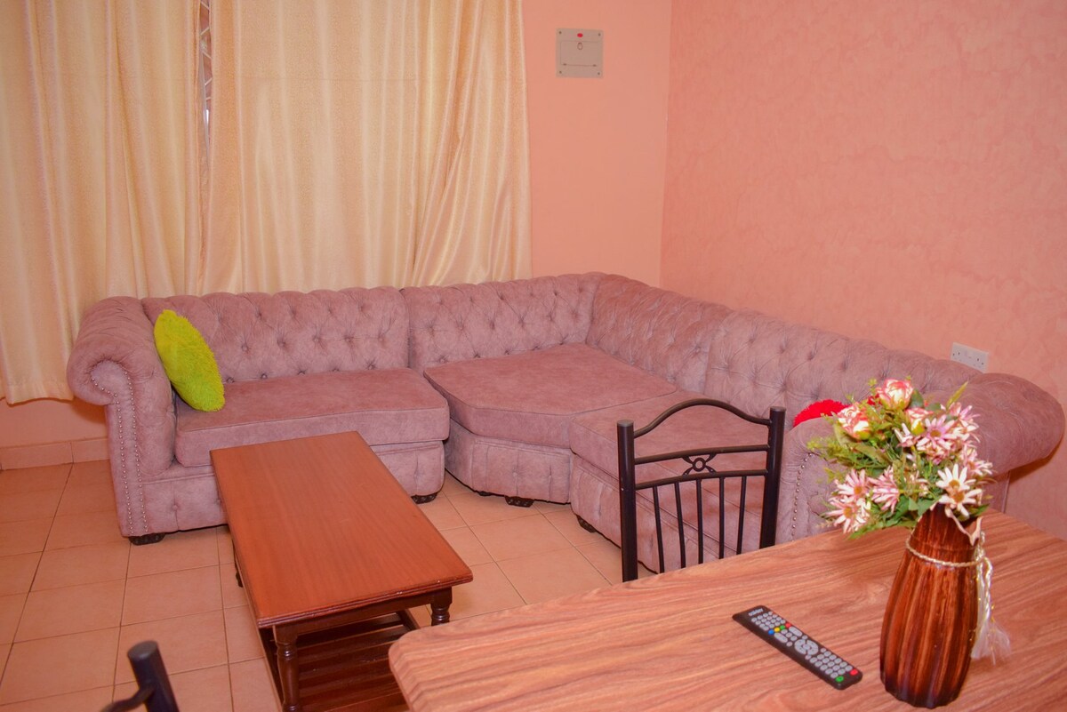 A spacious living area is displayed with a plush, tufted sectional sofa in soft pink. A wooden coffee table sits in front, complemented by a simple dining table with chairs nearby. Natural light filters through light-colored curtains, enhancing the comfortable atmosphere.