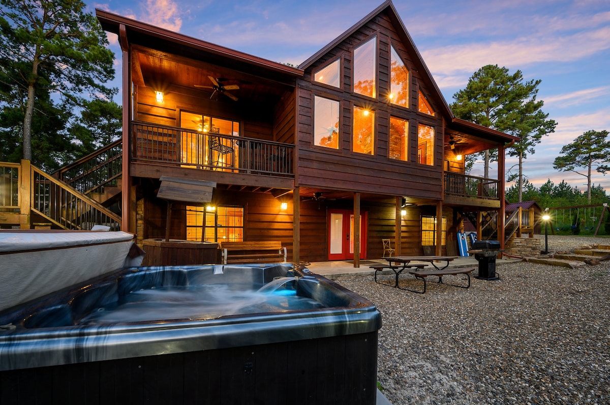 The exterior of the cabin showcases a striking design with large windows and wooden accents, set against a colorful sunset sky. A hot tub is visible in the foreground, positioned on a gravel patio, with picnic tables nearby for outdoor gatherings.