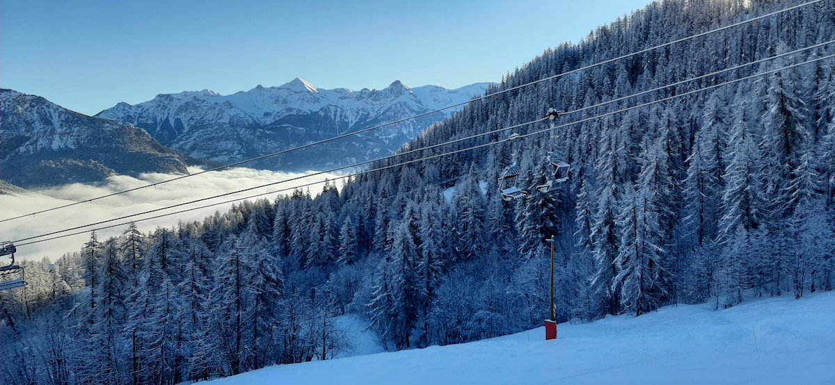 A snowy landscape is displayed, featuring tall trees blanketed in snow. The mountains in the distance are visible under clear blue skies. Ski lifts are seen in the foreground, indicating access to the slopes. The scene conveys a serene winter environment.