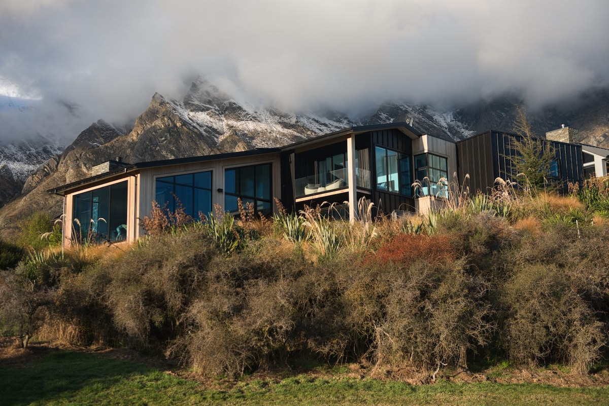 The contemporary architectural home is nestled against a backdrop of distinct mountain peaks, partially shrouded in clouds. Floor-to-ceiling windows line the exterior, providing abundant natural light and views of the surrounding landscape. Native vegetation and rustic terrain enhance the home's private setting.