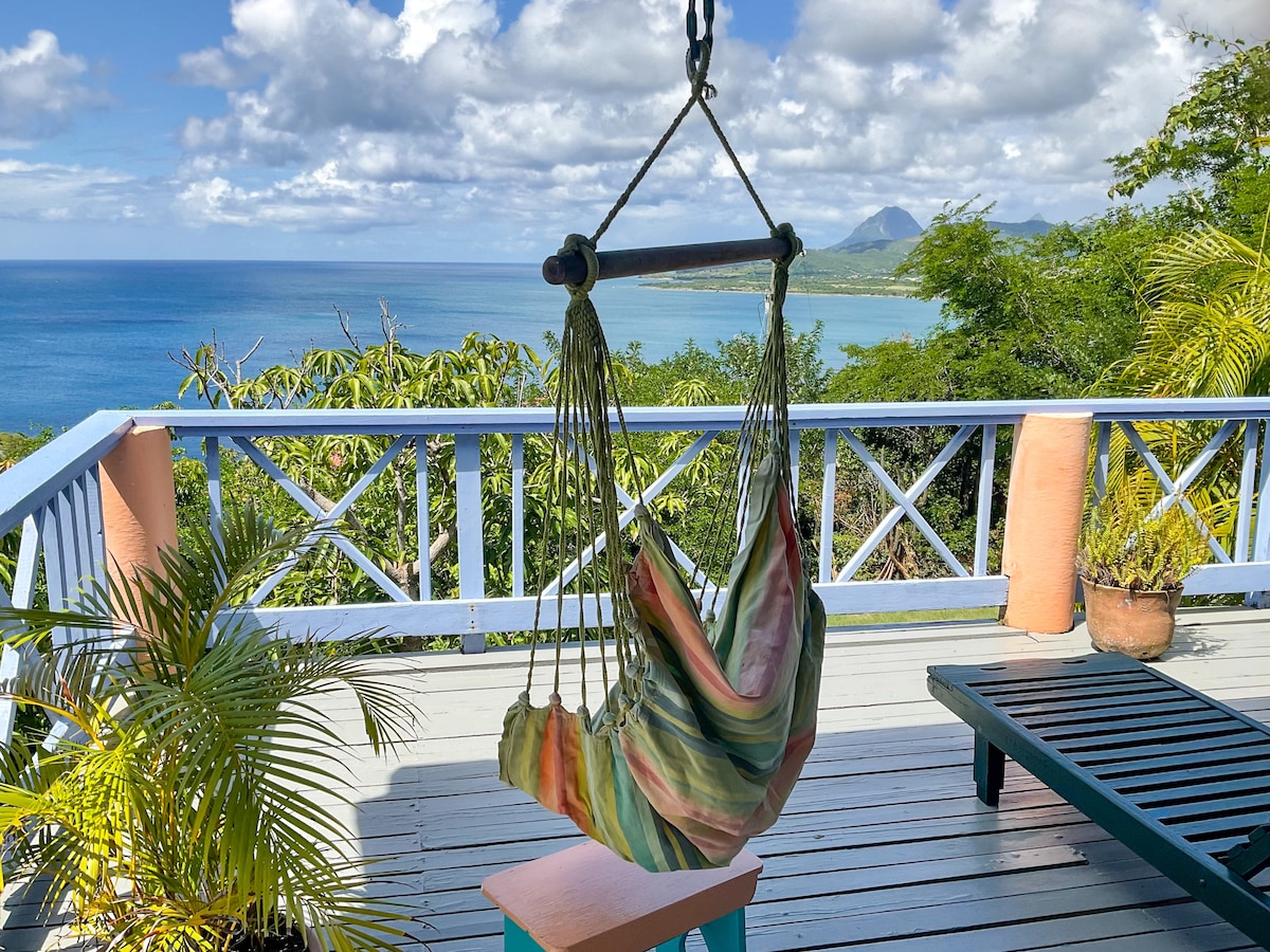 A colorful hammock is gently swaying under a bright sky, surrounded by lush greenery. The view includes the expansive Caribbean Sea and distant mountains, providing a tranquil setting for relaxation. A small potted plant and a lounging chair are positioned nearby on the wooden deck.
