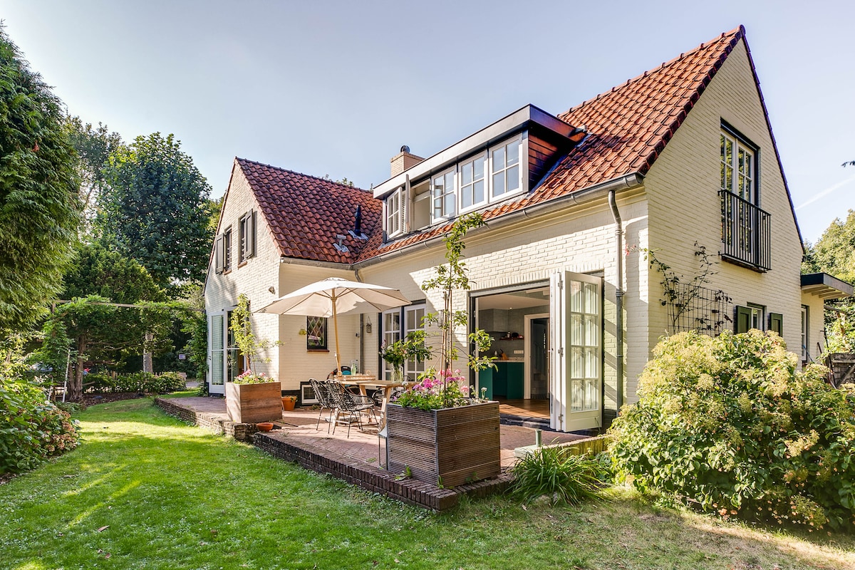 The spacious villa is surrounded by lush greenery, featuring a charming outdoor seating area with an umbrella. Large glass doors lead to an inviting interior, where natural light fills the open spaces. A well-maintained garden adds to the serene ambiance.