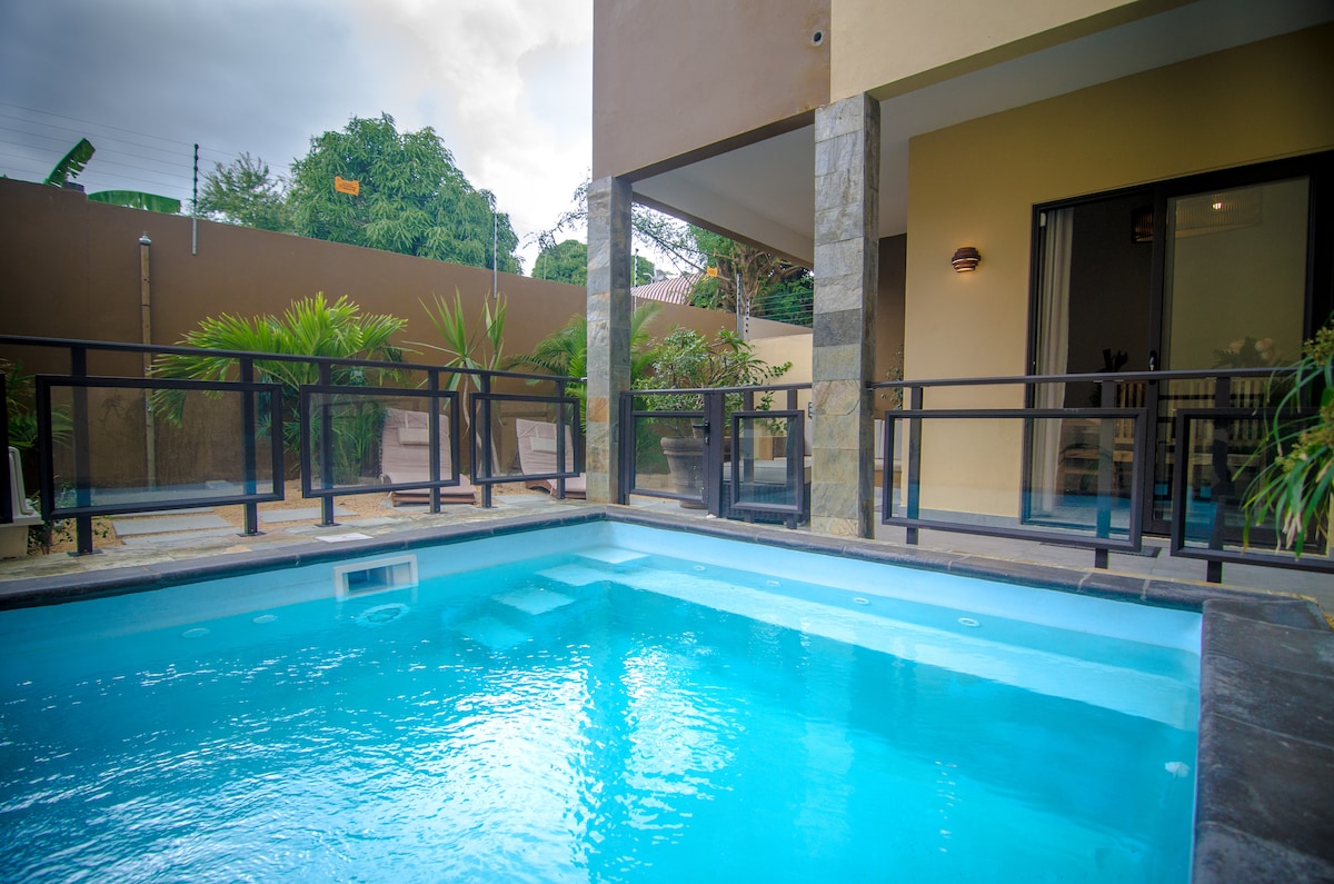 A clear, inviting pool is framed by a modern terrace with glass railings. Lush greenery surrounds the area, providing a serene backdrop. The building features large windows and a covered seating area, allowing for relaxation and views of the outdoor space.