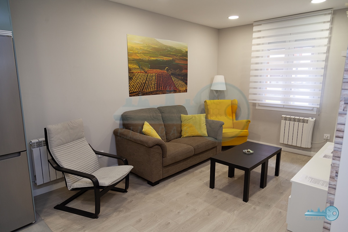 A modern living area is depicted, featuring a comfortable sofa alongside a bright yellow chair. A coffee table occupies the center space, while a large landscape artwork adorns the wall. Light streams in through partially covered windows, highlighting the room's neutral tones and sleek flooring.