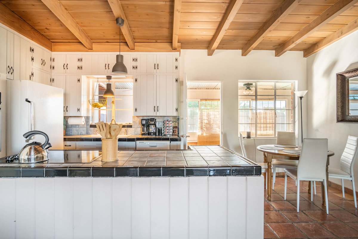 An open kitchen features wooden beams overhead and white cabinetry. A center island with a tiled countertop holds kitchen utensils. A stainless steel kettle and appliances are visible. Adjacent is a small dining area with a round table and four white chairs.