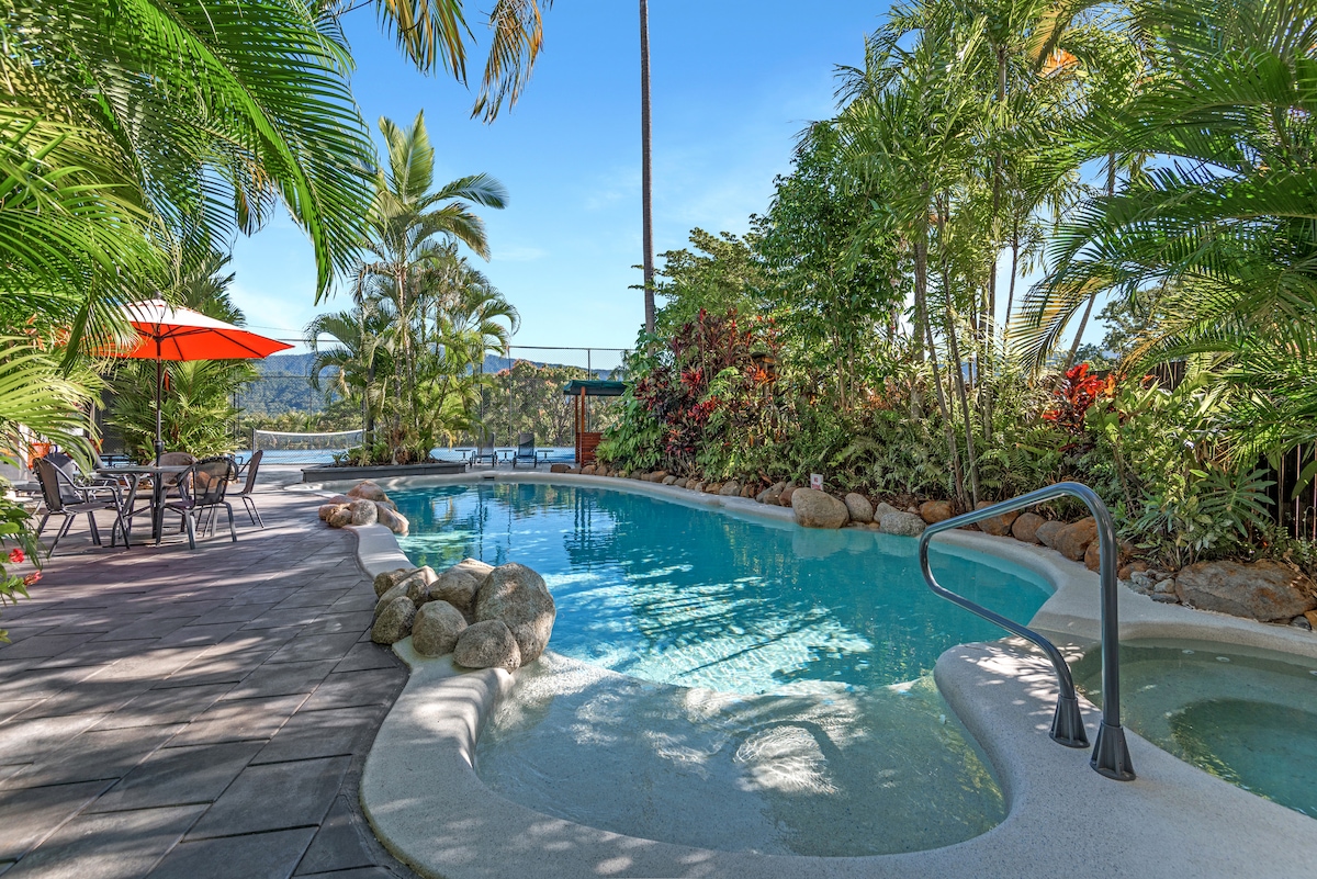 A tranquil pool area is surrounded by lush tropical plants and trees. A few lounge chairs and a shaded table with chairs are visible. The pool features a gentle curve, with smooth rocks incorporated into the landscaping, creating a serene outdoor space.