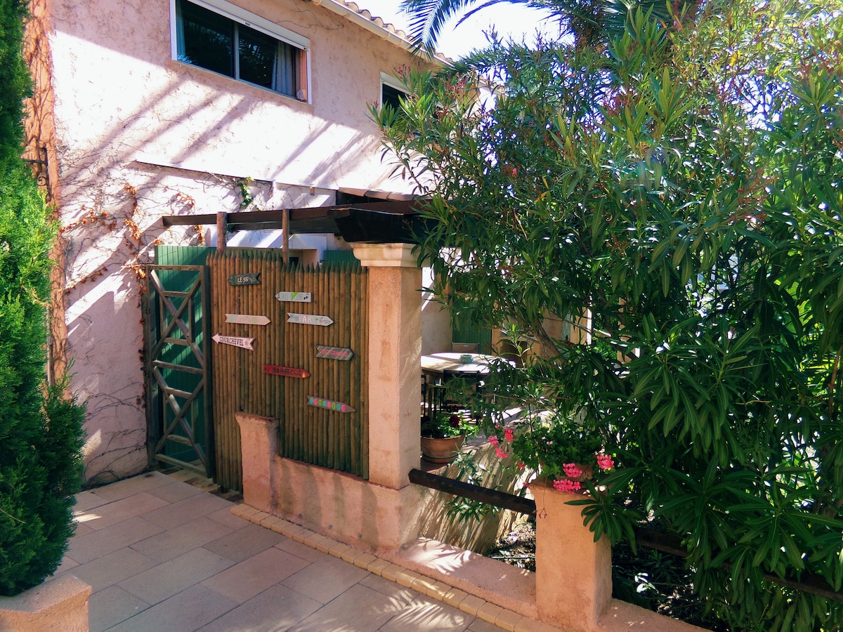 A private entrance is framed by vibrant greenery and decorative fencing. Light filters through the trees, casting gentle shadows on the stone pathway, which leads to a cozy outdoor area surrounded by lush plants and colorful flowers.