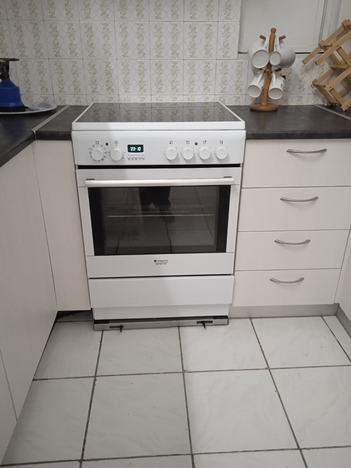 A white kitchen stove is positioned against a light-colored wall with decorative tiles. The stove features an oven with a glass door and control knobs on the front. Nearby, a simple dish rack and a set of hanging mugs are visible, complementing the clean, functional design.