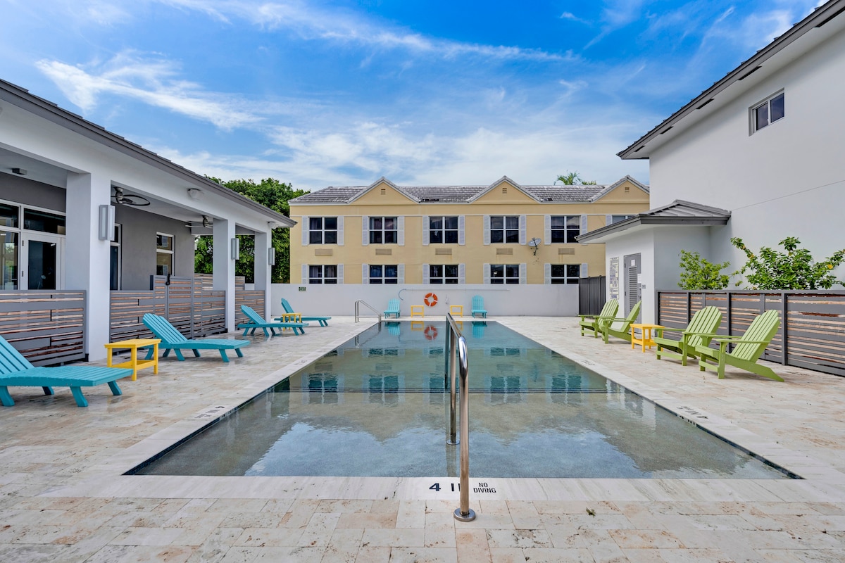 A modern pool area is framed by lounge chairs in vibrant turquoise and green. The pool is lined with a light stone surface, reflecting the clear sky and surrounding buildings. A lifebuoy hangs on the wall near the shallow end of the pool, offering safety assurance.