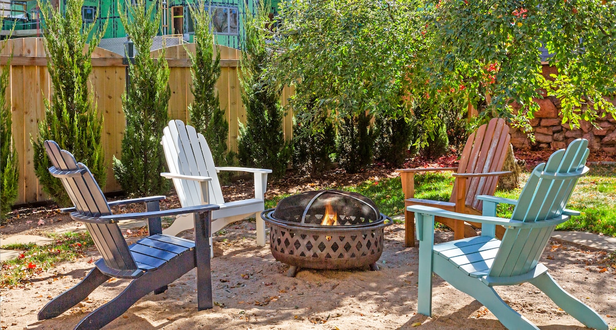 A circular fire pit is surrounded by four Adirondack chairs in varied colors. The sandy ground is framed by green grass and a backdrop of tall, leafy plants, providing a cozy setting for outdoor gatherings.