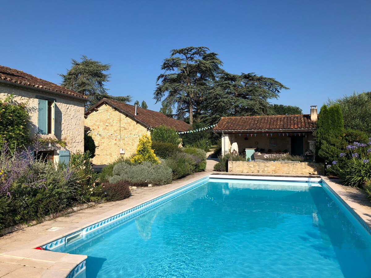 A clear blue pool is surrounded by landscaped gardens, featuring flowering plants and fragrant shrubs. The farmhouse buildings, made of stone and featuring terracotta roofs, are visible in the background against a cloudless sky. The outdoor kitchen and BBQ area are also nearby.