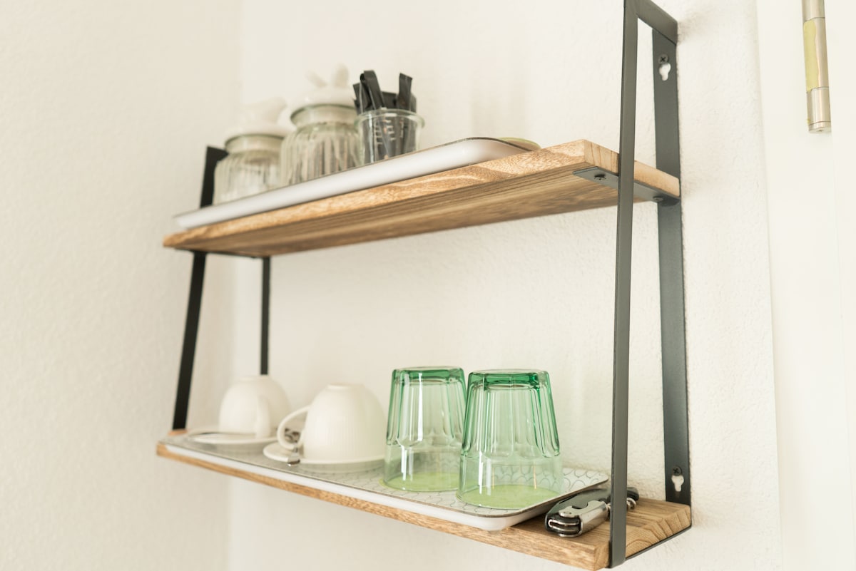 Two open shelving units display a variety of kitchen items. The top shelf features glass jars, utensils, and a tray. The bottom shelf holds four white cups and two green glasses on a patterned tray, providing convenient access to dining essentials.