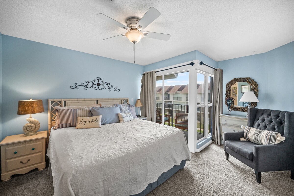 A serene bedroom features a spacious bed with a decorative light-colored quilt and two bedside lamps. A cozy armchair sits in one corner, while large sliding glass doors open to a private balcony, allowing natural light to fill the room.