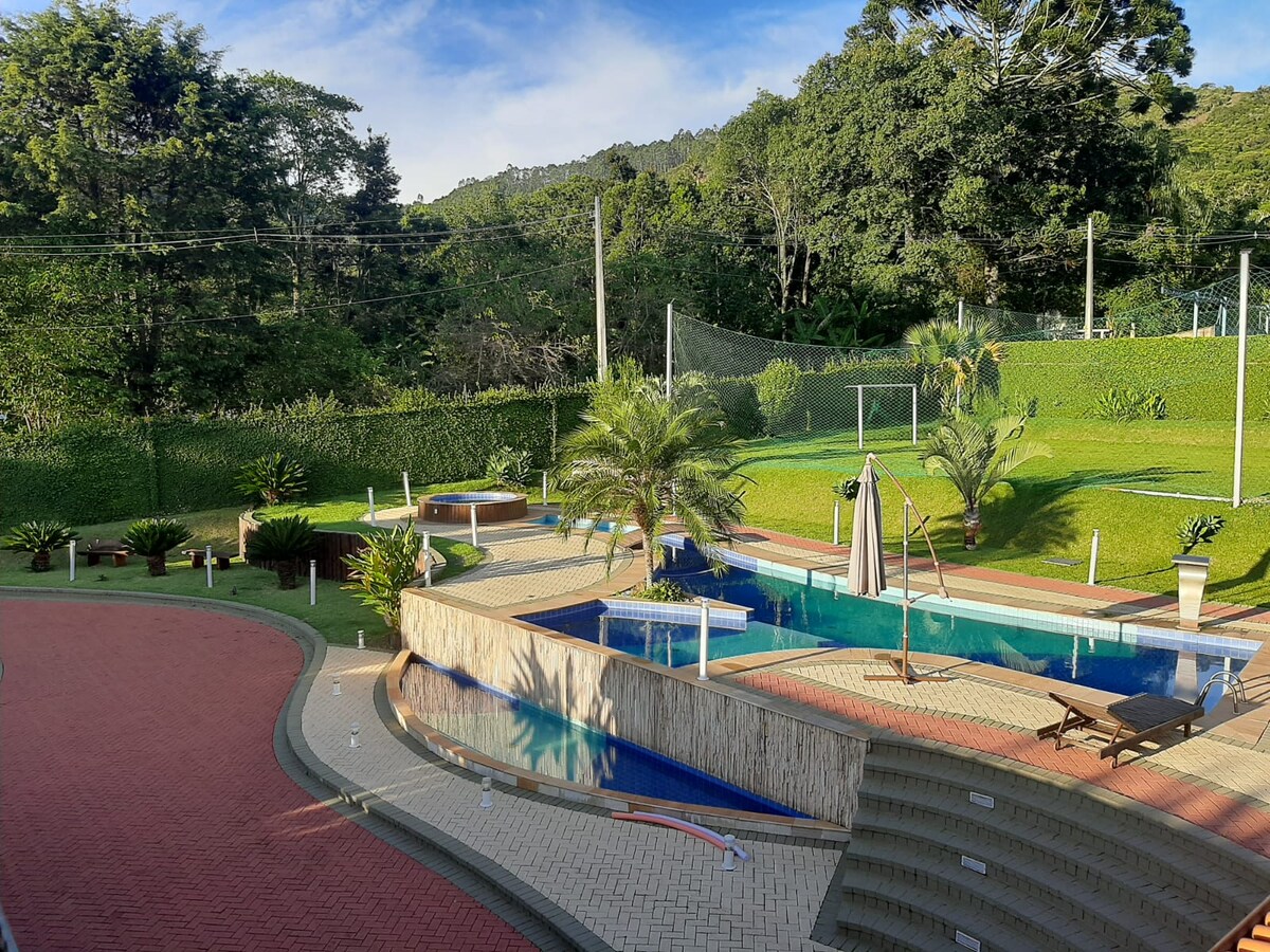 A spacious outdoor area features multiple swimming pools surrounded by lush greenery. Deck chairs and a swing are positioned nearby, offering a comfortable relaxation space. A manicured lawn extends into the background, showcasing trees and a sports field framed by tall hedges.