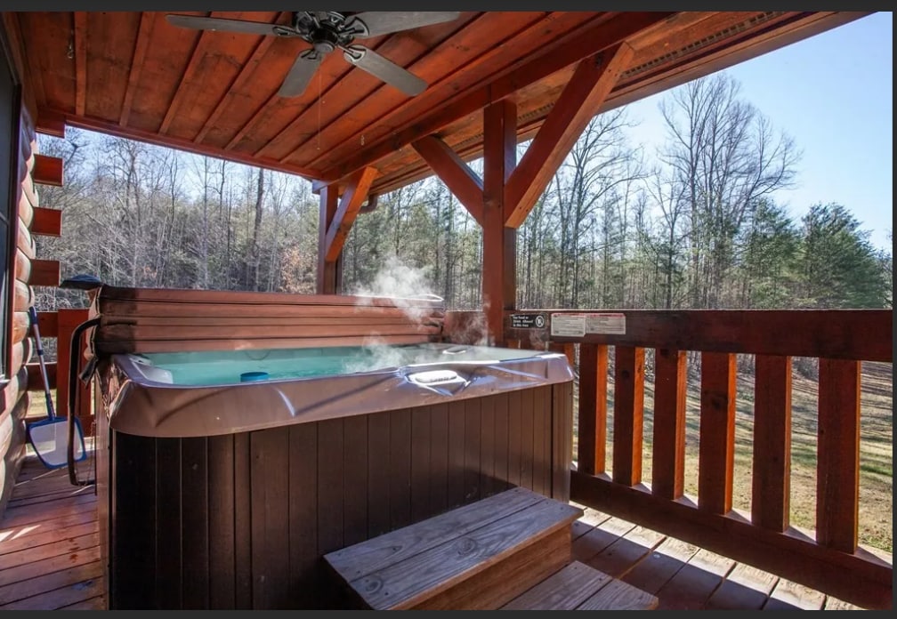 A luxurious hot tub is situated on a covered porch, emanating steam in the cool air. The wooden structure features a ceiling fan overhead and offers a peaceful view of the surrounding trees and expansive yard, enhancing the outdoor experience.