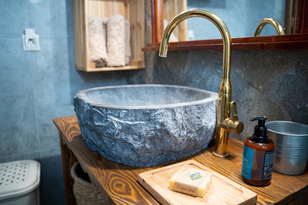 A stone basin provides a natural element to the bathroom, accompanied by a sleek gold faucet. Wooden shelving displays neatly arranged towels, while a small wooden tray holds a bar of soap. Textured tiles in shades of gray create a serene ambiance.