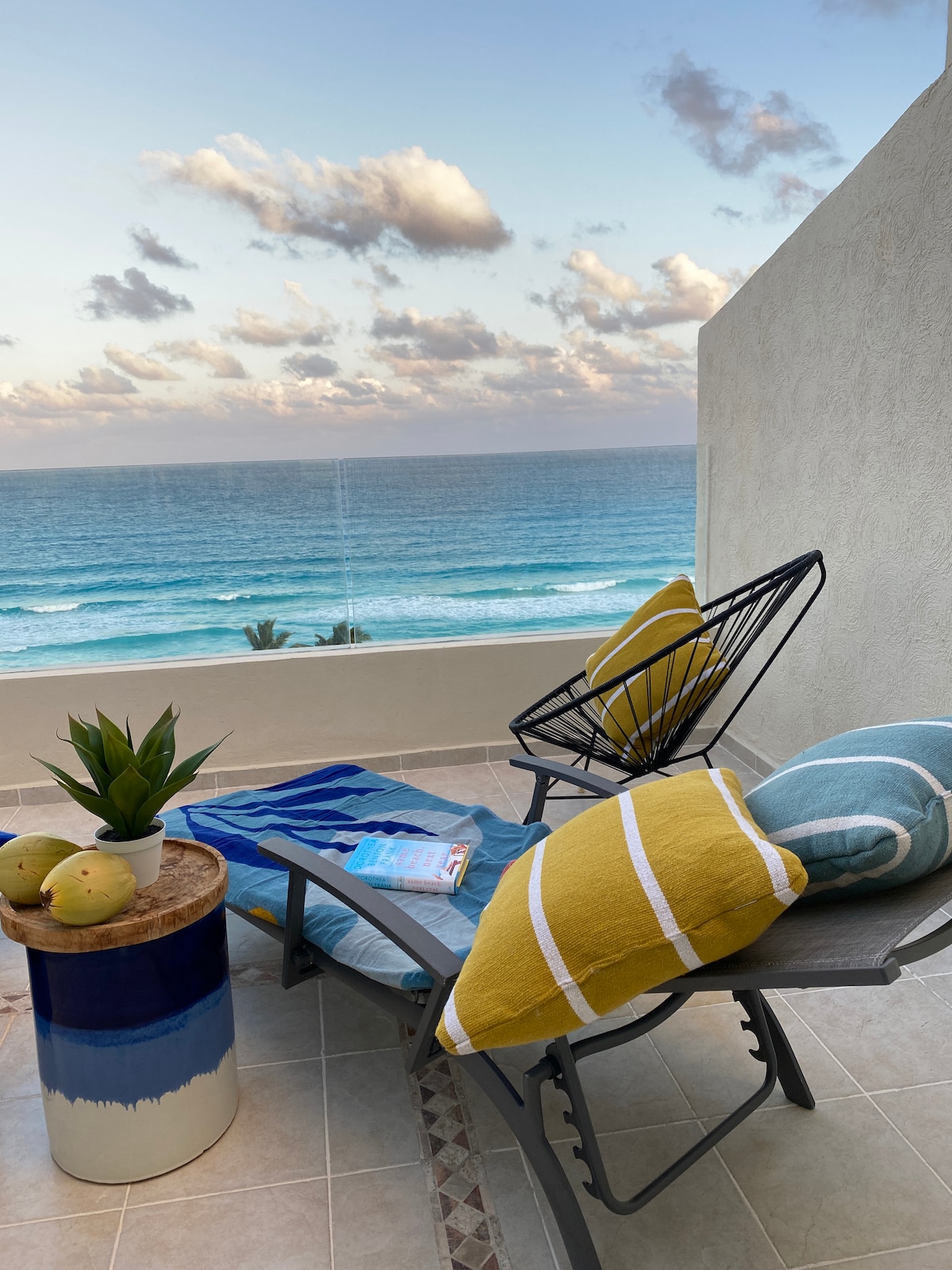 A private balcony offers a tranquil setting with views of the ocean. A comfortable lounge chair, accented with colorful cushions, is paired with a small wooden table displaying decorative elements, while a potted plant adds a touch of greenery. Soft lighting is present as the day transitions into evening.