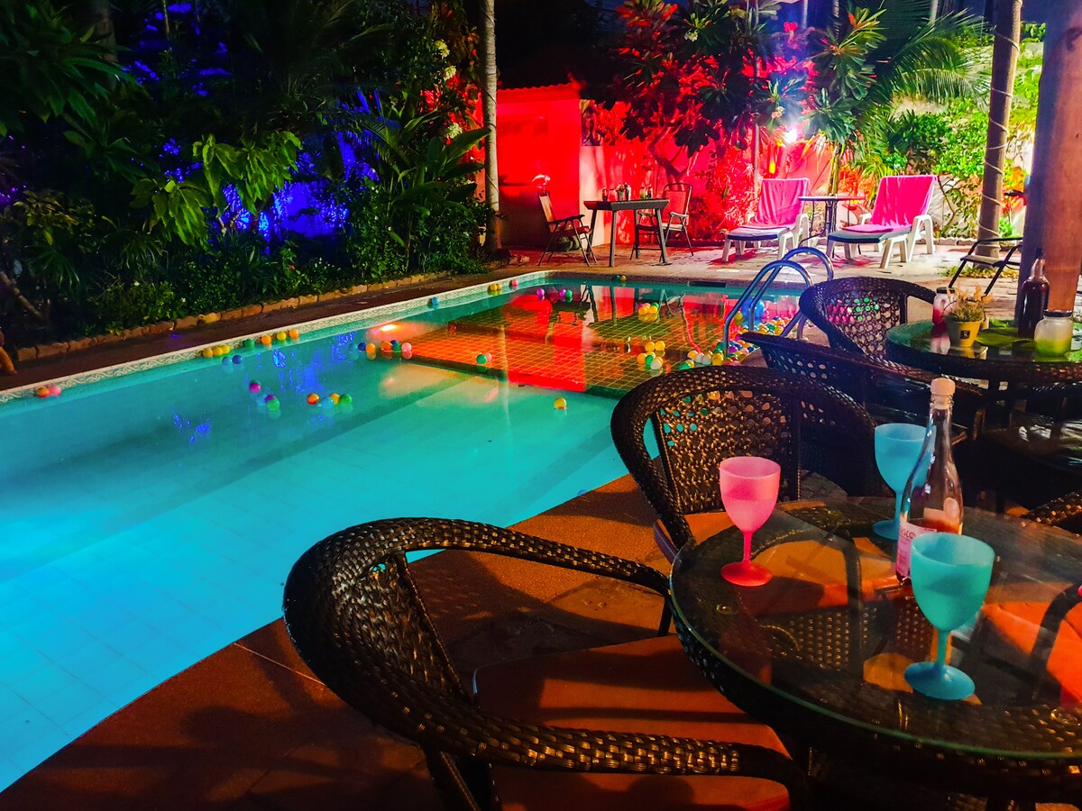 A large private pool is surrounded by tropical gardens, featuring vibrant lighting in shades of blue and red. Colorful floating decorations dot the water, while stylish wicker tables with clear glasses sit at the edge, providing a relaxed atmosphere for guests.